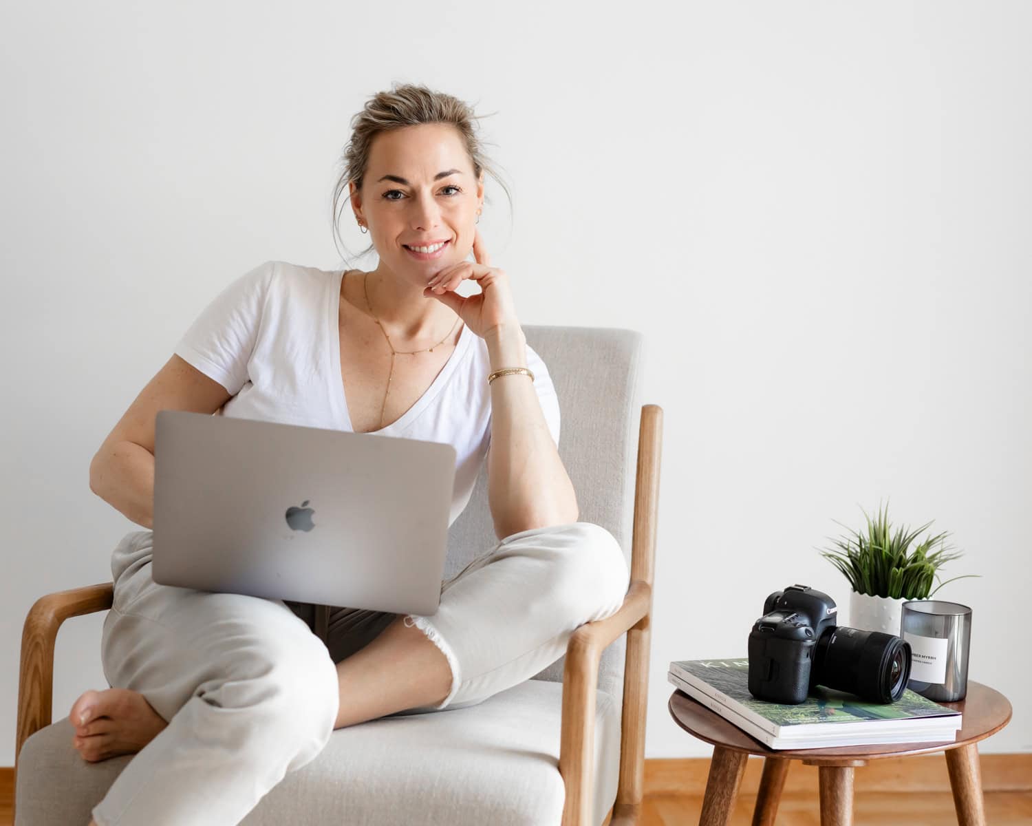 female photographer entrepreneur branding image of her sitting on a chair with a computer on the lap and a camera on the side table next to her